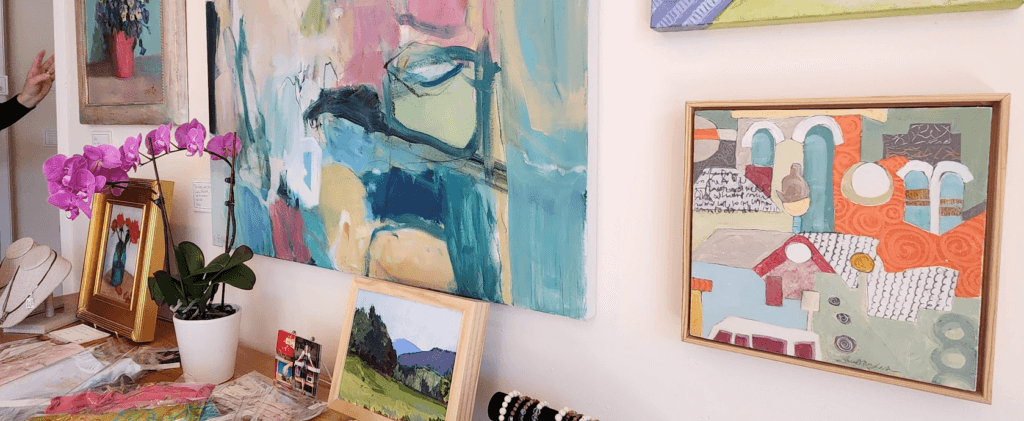 Rutledge Gallery Art work for sale-wide - Experience Camden, South Carolina A wall displays colorful abstract and landscape paintings. A table below holds a purple orchid, beaded jewelry, necklaces on stands, and a small framed landscape photo. A persons hand is partially visible to the left.