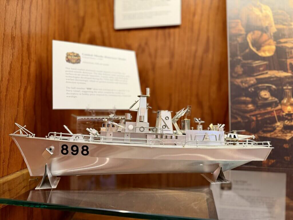 Metal model battleship with the number 898 on its bow, displayed on a glass shelf against a wooden backdrop.