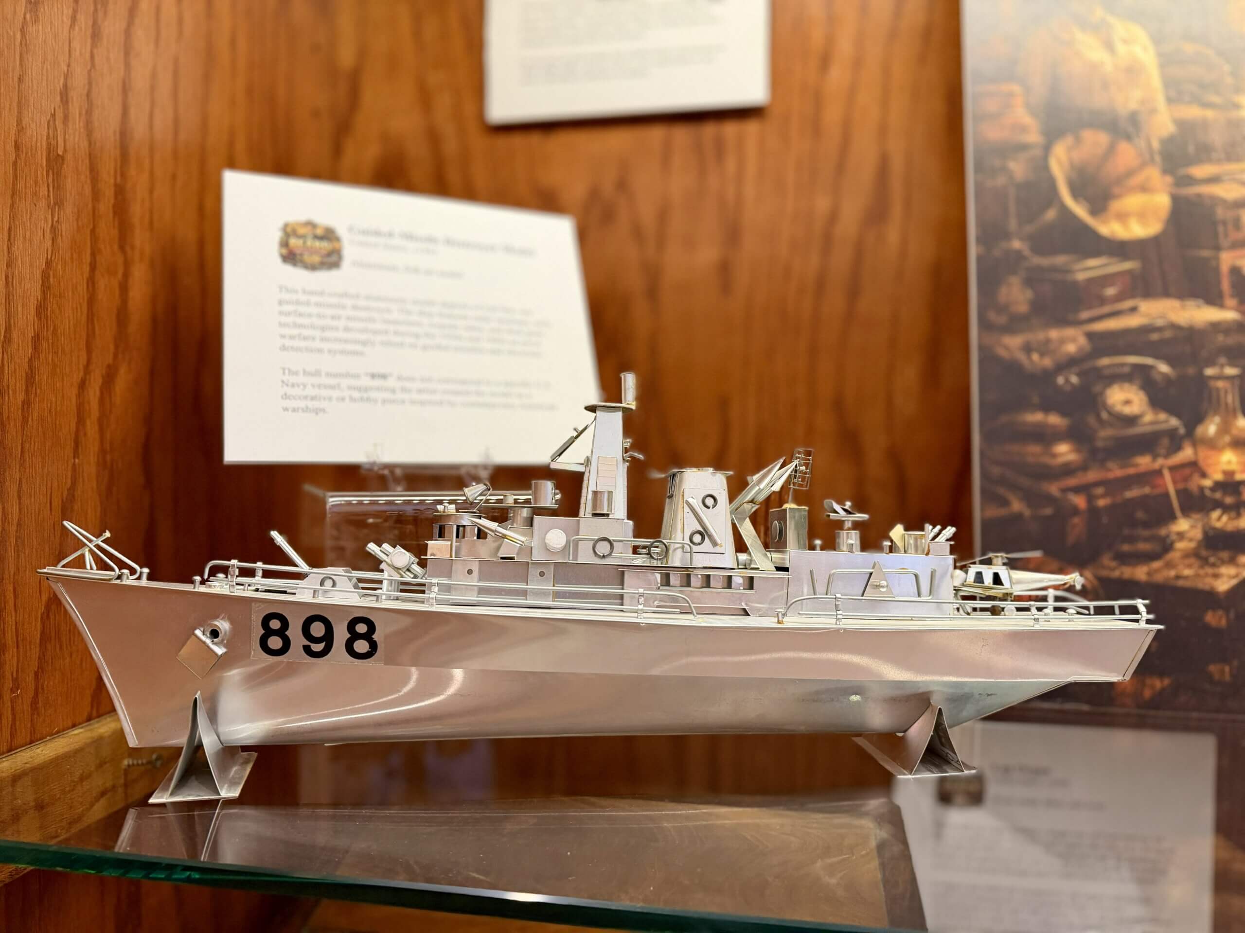 IMG_4400 Metal model battleship with the number 898 on its bow, displayed on a glass shelf against a wooden backdrop.