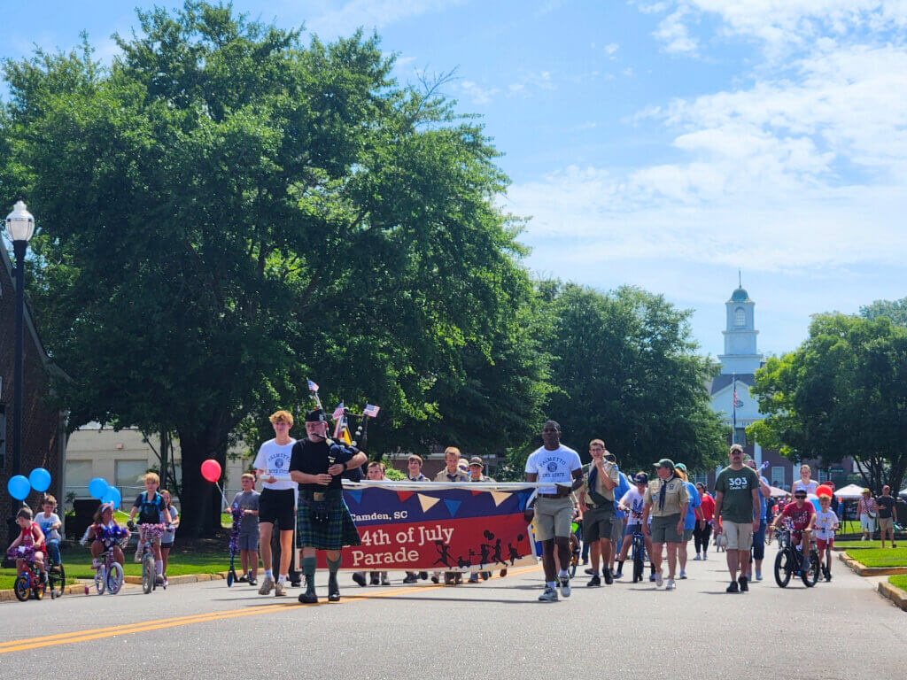 0658a28b-90ed-4d1c-b98f-a0fe9b15b7ba - Experience Camden, South Carolina Parade marching down a sunny street with a red banner, families on bicycles and walkers, patriotic balloons visible on sides.