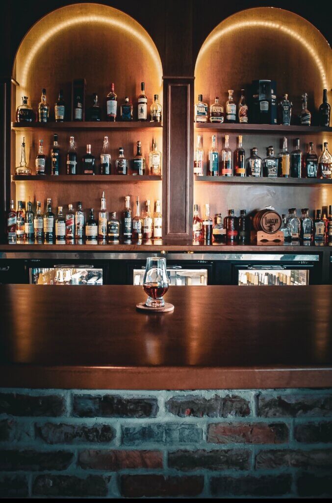 Screenshot - Experience Camden, South Carolina Bar interior with backlit bottles on shelves and a single glass of whiskey centered on a dark wooden counter.