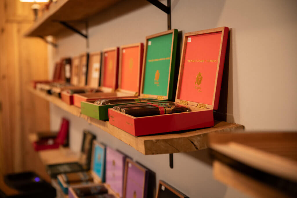Copy of Cigar Club-28 - Experience Camden, South Carolina Colorful open wooden boxes with bright red, green, and pink packaging on a shelf in a cozy shop (blurred foreground).