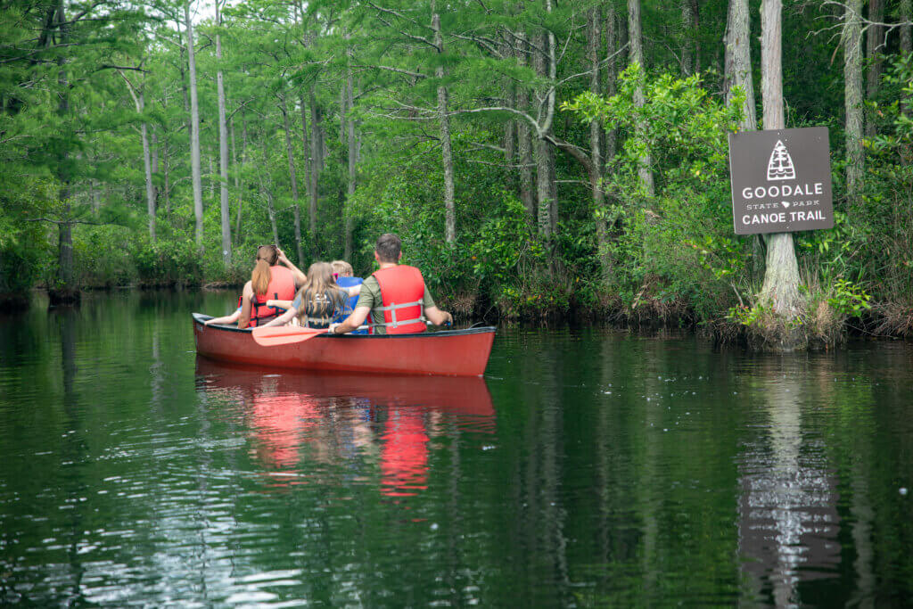 Copy of Day 3-183 1 - Experience Camden, South Carolina Group of people in a red canoe wearing life jackets paddling along a narrow, green forested river; a Goodale State Park Canoe Trail sign is on the bank.