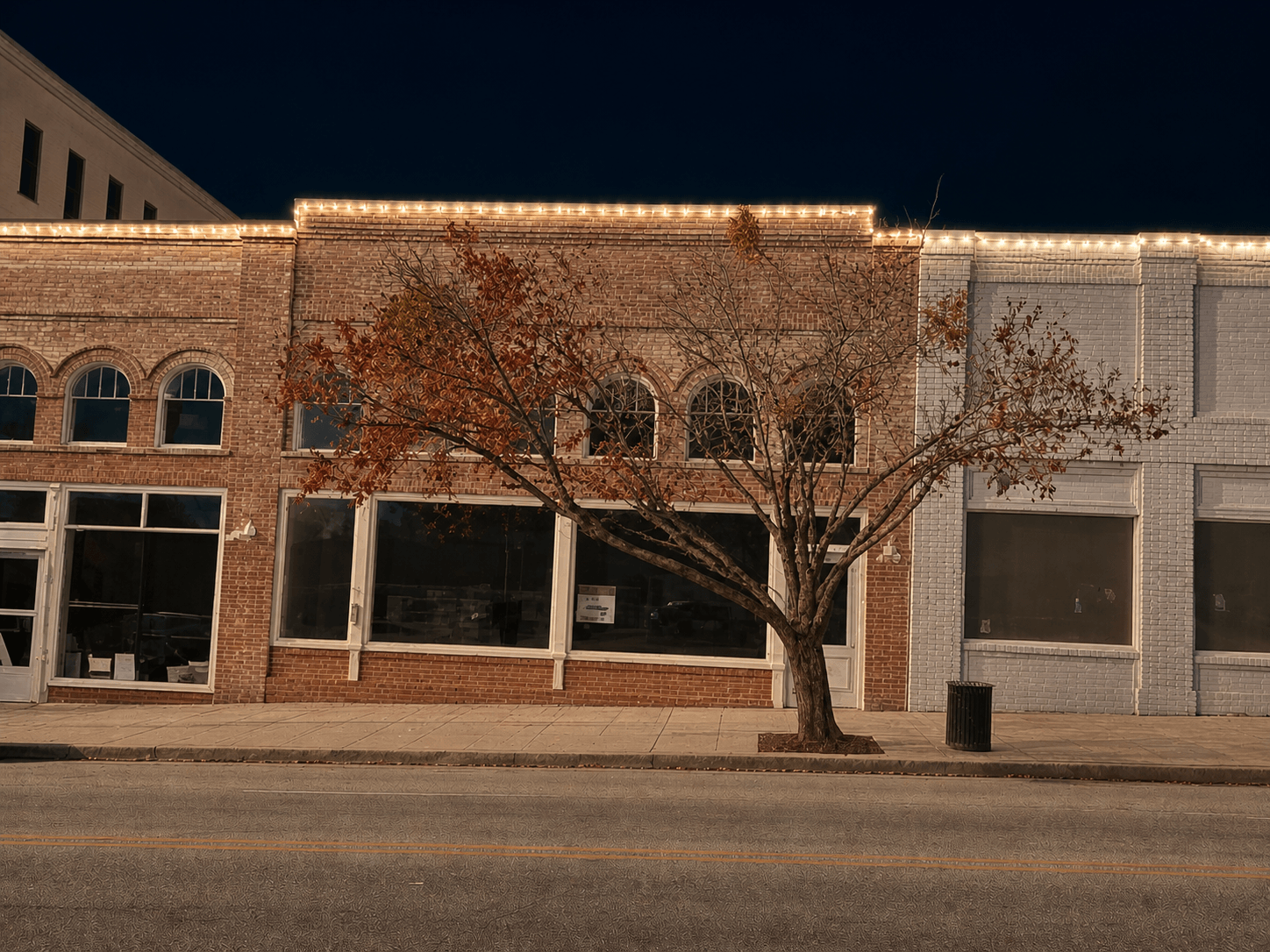 DT Camden Storefront LED Lighting_ Nighttime street view of brick storefronts with arched windows and a leafless tree in front, string lights along the roof.