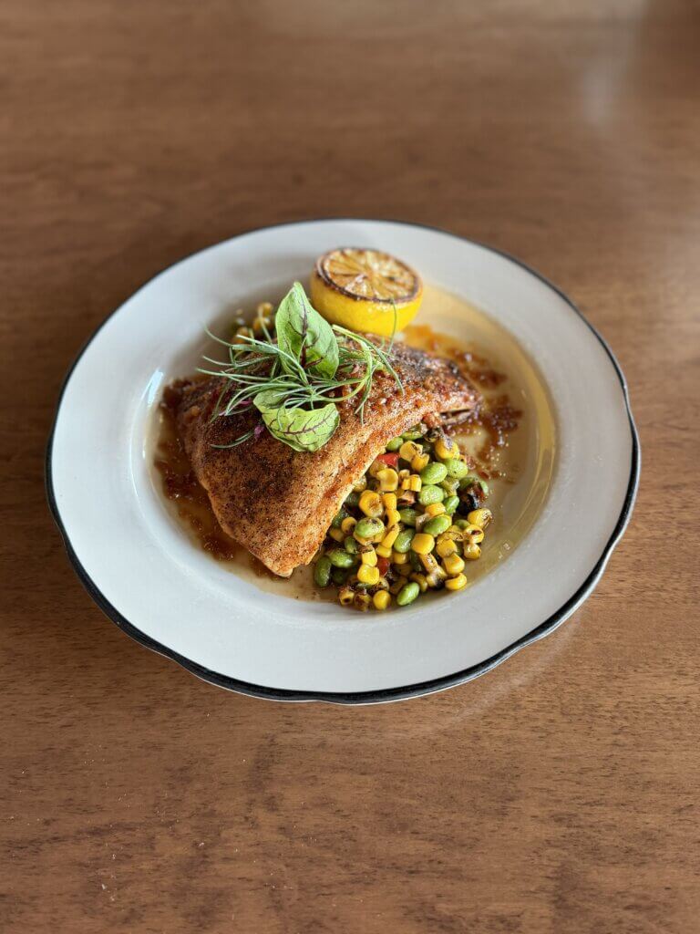 IMG_4437 - Experience Camden, South Carolina Seared fish fillet on a bed of corn and edamame, garnished with microgreens and a grilled lemon half on a white plate.