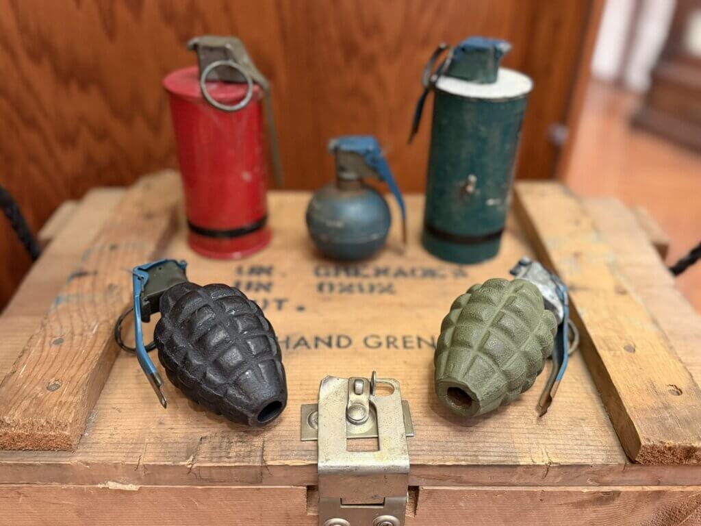 IMG_4606 - Experience Camden, South Carolina Antique military grenades and fuzes on a wooden crate, red and green metal cans with worn paint and safety pins visible.