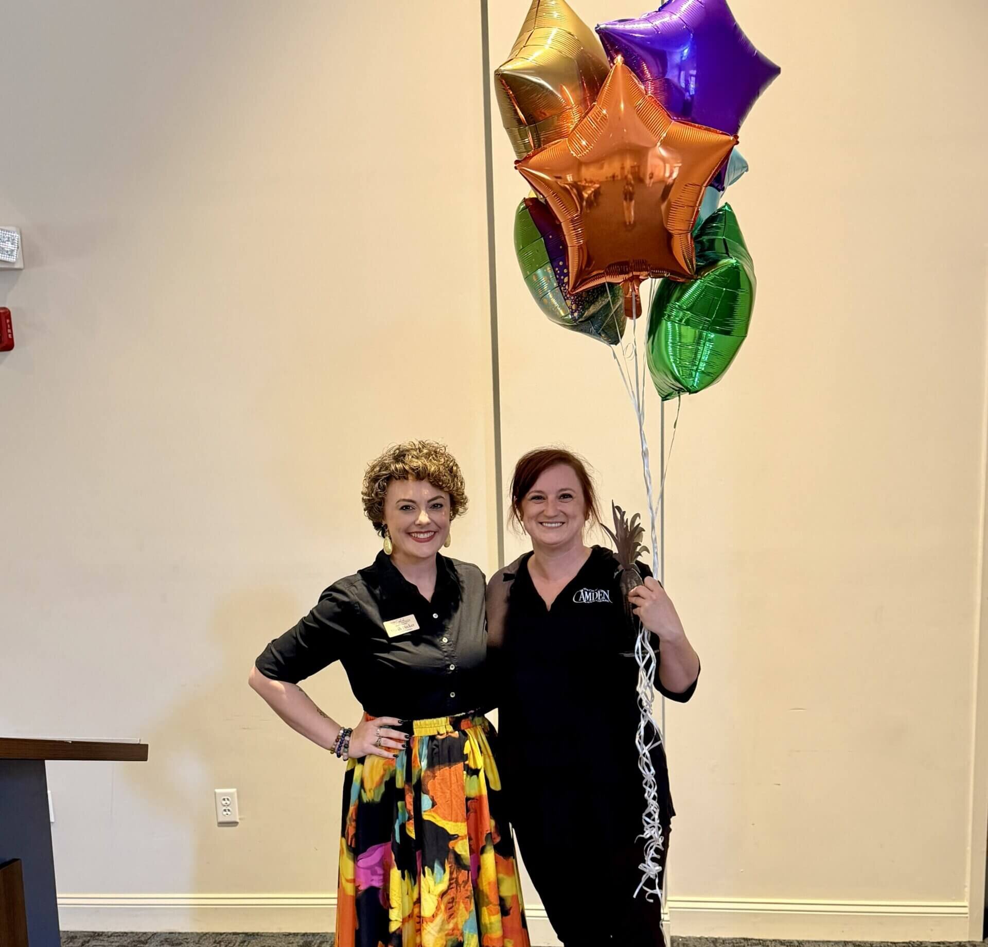 IMG_4712 Two women pose indoors with colorful star-shaped balloons; one wears a black top and a bright floral skirt, smiling at the camera.