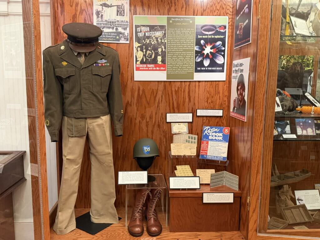 Military uniform on a mannequin with a peaked cap and ribbons, displayed beside WWII-era posters on a wood-paneled wall.