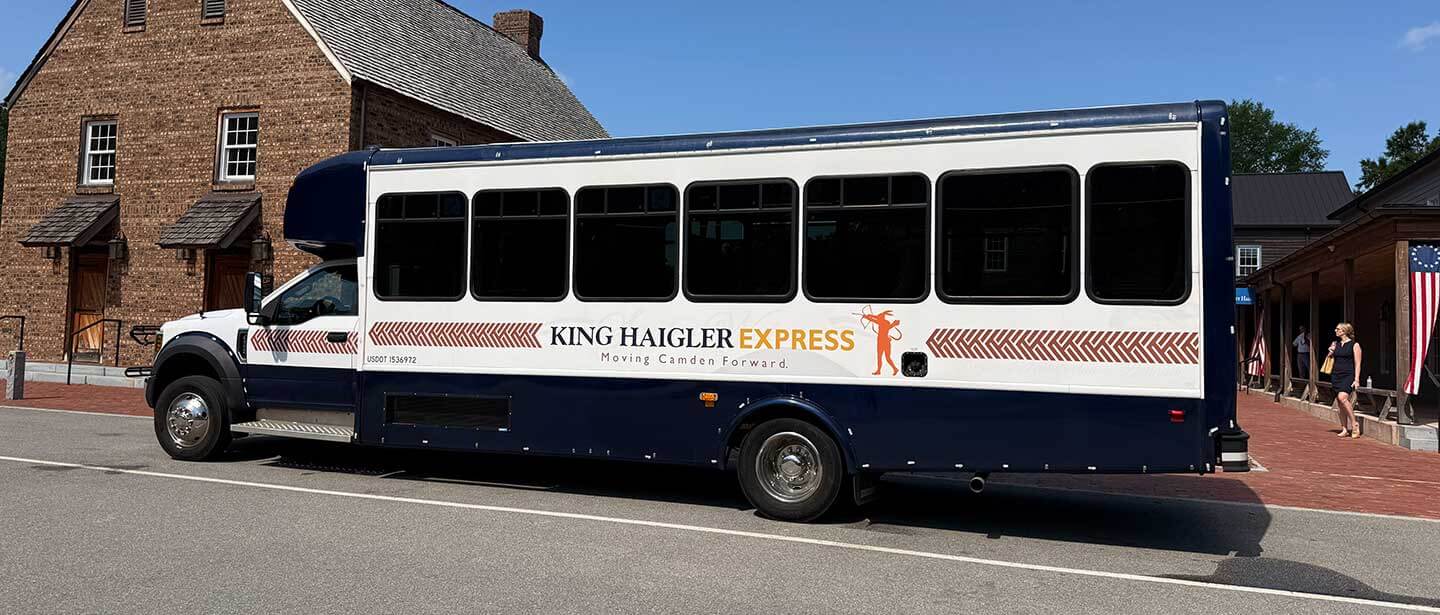 IMG_5278 Side view of a navy and white King Haigler Express shuttle bus parked on a sunny street beside a brick building, with a person near an American flag in the background.