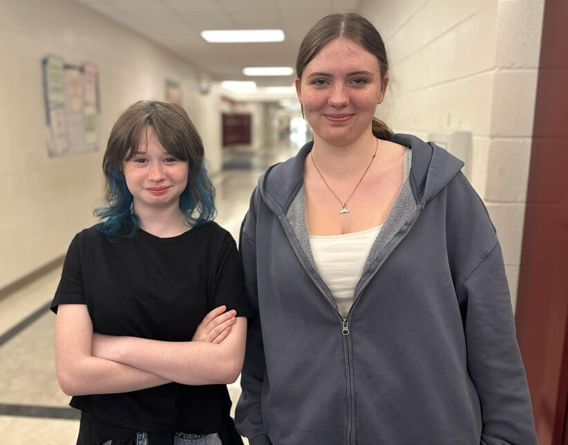 Student Art Work Winners 002 Two teenage girls stand in a school hallway, smiling; one has blue-tipped hair and a black T-shirt, the other wears a gray zip hoodie and white top.
