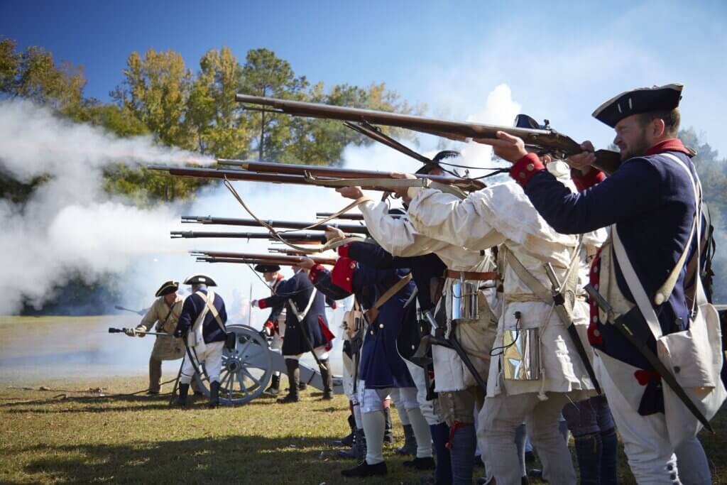 Line of soldiers in blue and white 18th-century uniforms firing muskets in a smoke-filled outdoor reenactment scene