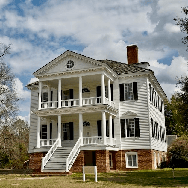 image - Experience Camden, South Carolina White two-story house with a brick foundation, large columned porches, and black shutters under a cloudy sky outside a yard.