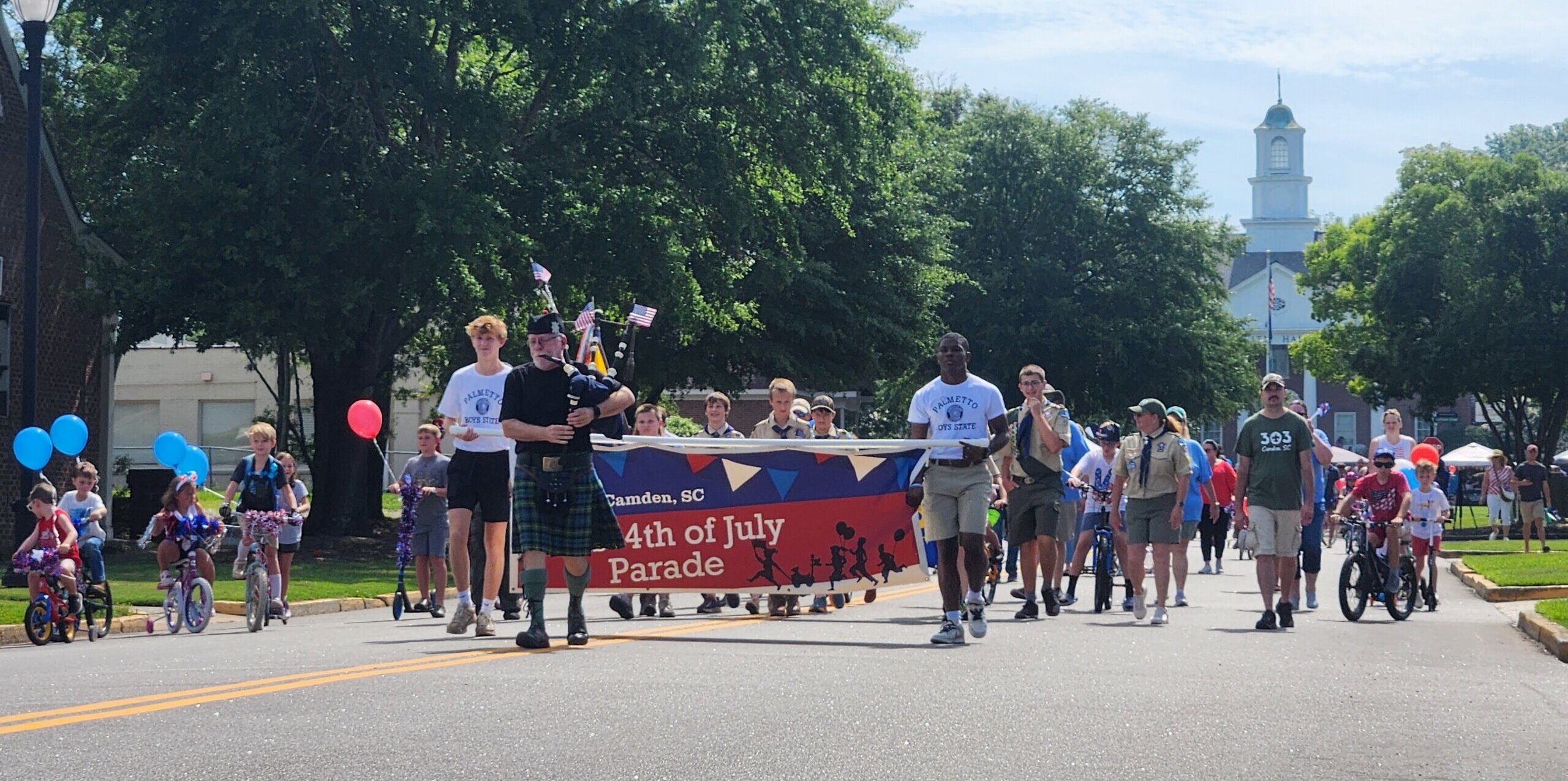 2025 kids parade People march in a July 4th parade down a sunny street, carrying a large red banner that reads '4th of July Parade' with Camden, SC visible; spectators and balloons along the sides.