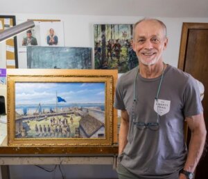 IMG_2635 - Experience Camden, South Carolina A smiling older man stands in a studio beside a framed painting depicting a historical battle scene with soldiers, a blue flag, and wooden buildings. Various artworks and supplies are visible in the background.