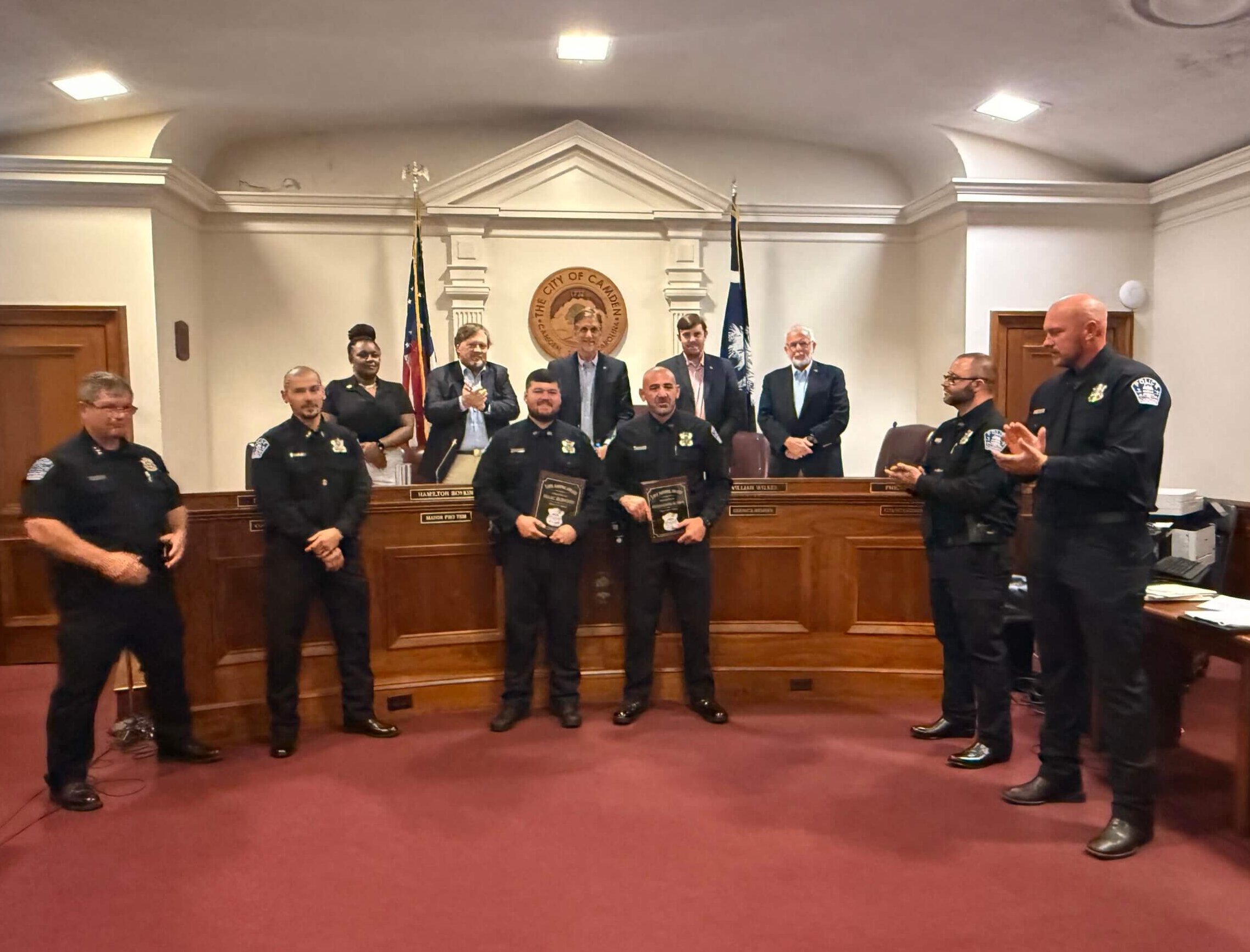 IMG_6046 Police officers and officials posing for an awards ceremony in a city council chamber, with the city seal and flags in the background.
