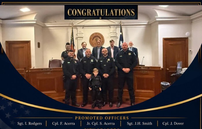 Screenshot 2026-06-03 085000 Group of police officers posing with a child in front of a dais under a 'CONGRATULATIONS' banner on a ceremony stage.