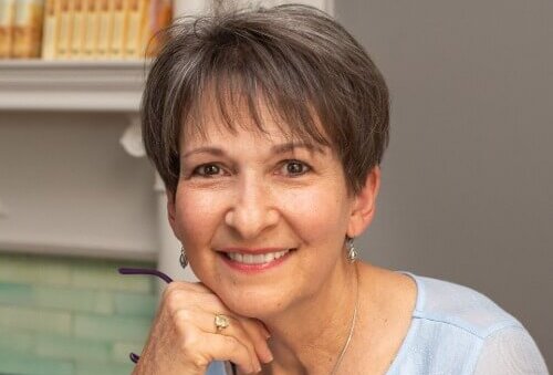 donna everhart.jpg - Experience Camden, South Carolina Smiling woman with short hair resting her chin on her hand, holding glasses, in a light blue sweater in a cozy indoor setting with a bookshelf behind her.
