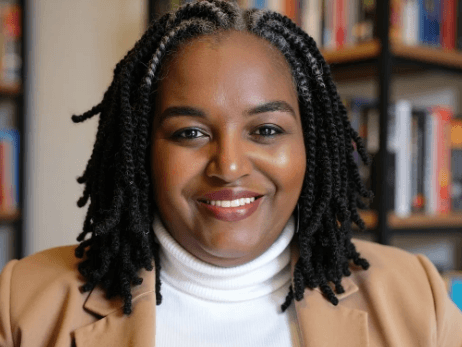 2026_07_01_17_27_19.png - Experience Camden, South Carolina A woman with shoulder-length twists, wearing a tan blazer and white turtleneck, smiles in front of bookshelves filled with books.