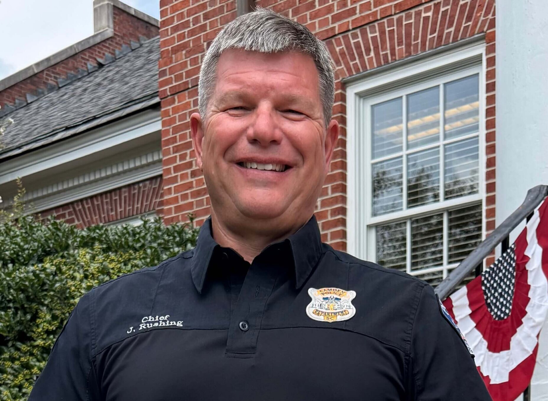 Chief Rushing Graduates FBI NCC Smiling man in a black uniform shirt stands outside, holding an FBI certificate while posing on brick steps decorated with patriotic bunting.