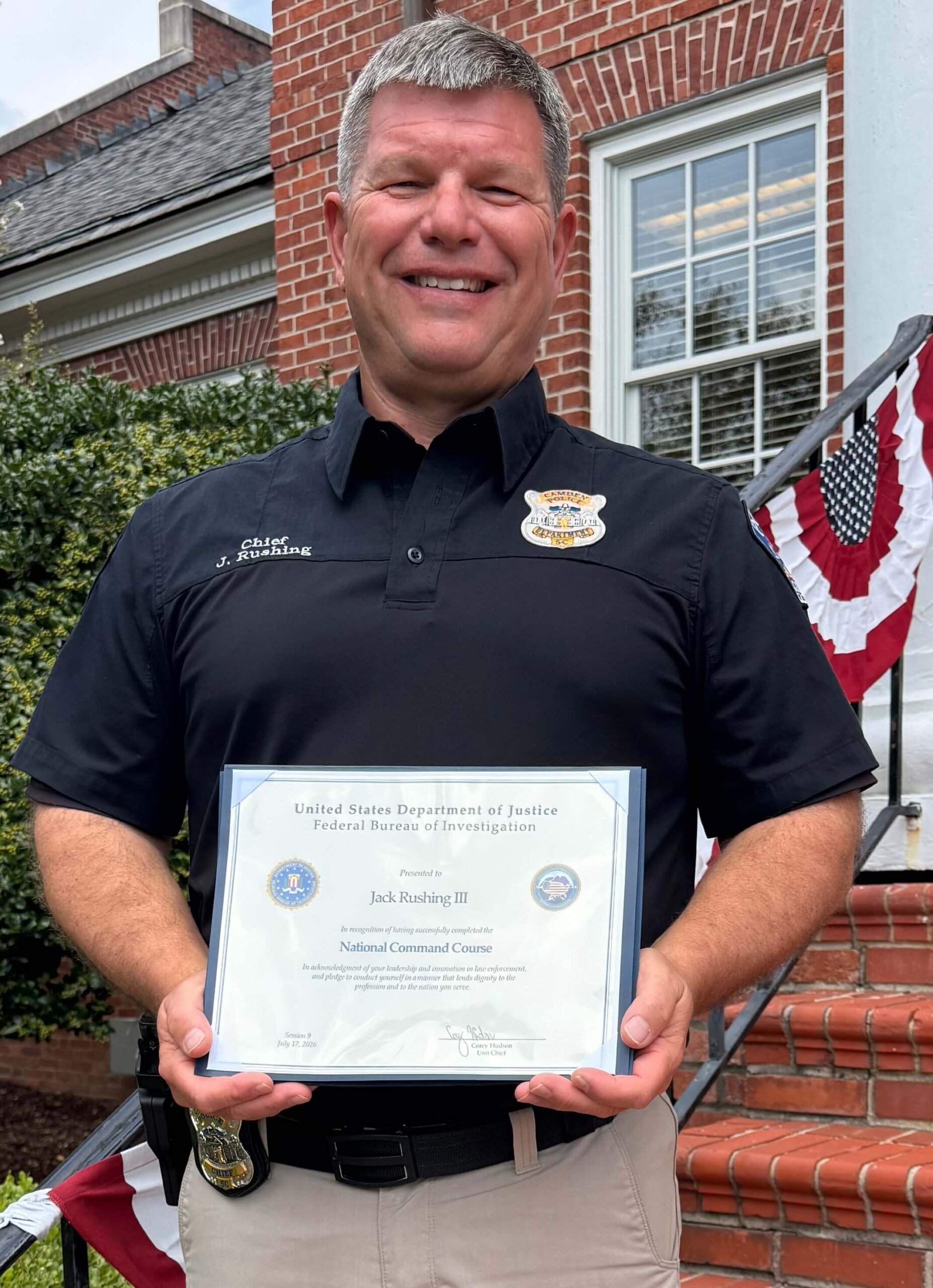 Chief Rushing Graduates FBI NCC Smiling man in a black uniform shirt stands outside, holding an FBI certificate while posing on brick steps decorated with patriotic bunting.