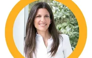 Jacqueline-Friedland.webp - Experience Camden, South Carolina A woman with long brown hair, wearing a white blouse, smiles at the camera. She stands in front of a window with greenery outside. The photo is framed by a thick yellow circle.