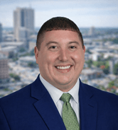 Matts New Headshot – USE THIS ONE A man in a blue suit, white shirt, and green tie smiles in front of a blurred cityscape background.