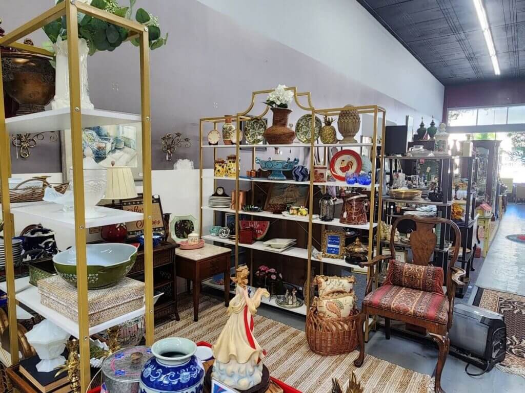antique - vc - Experience Camden, South Carolina Shelves and tables filled with assorted vintage items, ceramics, vases, figurines, lamps, and decorative objects in a well-lit antique store with a rug and chairs in the foreground.