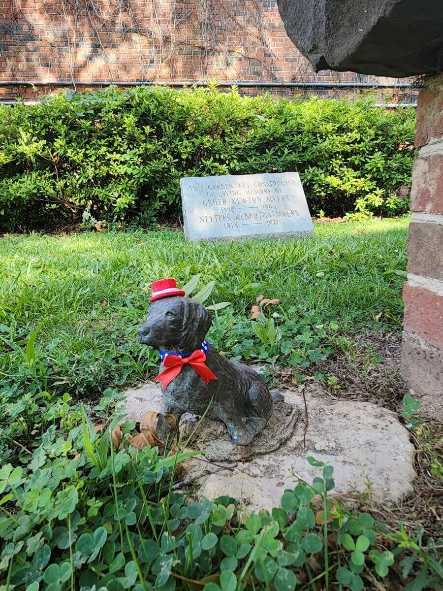 boykin 250th - Experience Camden, South Carolina A small dog statue with a red, white, and blue hat and bow sits on a stone in a grassy yard. In the background, a memorial plaque is partially visible among green bushes.