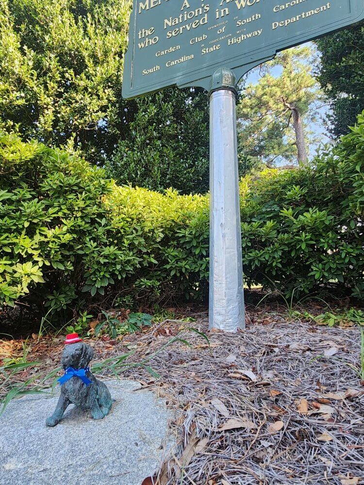 boykin2 - Experience Camden, South Carolina A small dog statue wearing a red Santa hat and blue scarf sits on the ground near a memorial sign surrounded by bushes and trees.