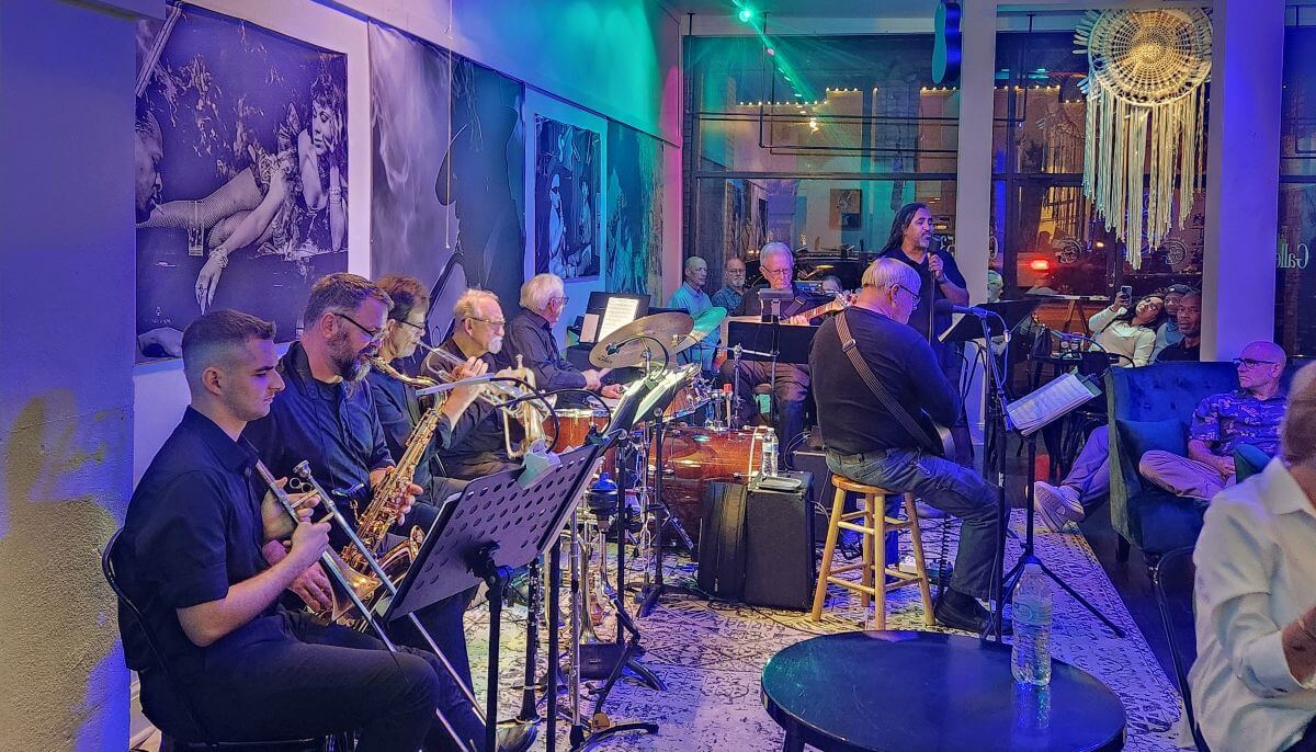 gallery 537 - Experience Camden, South Carolina A jazz band performs live in a cozy, dimly lit lounge with blue and green lights. Musicians play various instruments, and a singer stands at a microphone. People watch and relax at tables along the side. Large art decorates the walls.
