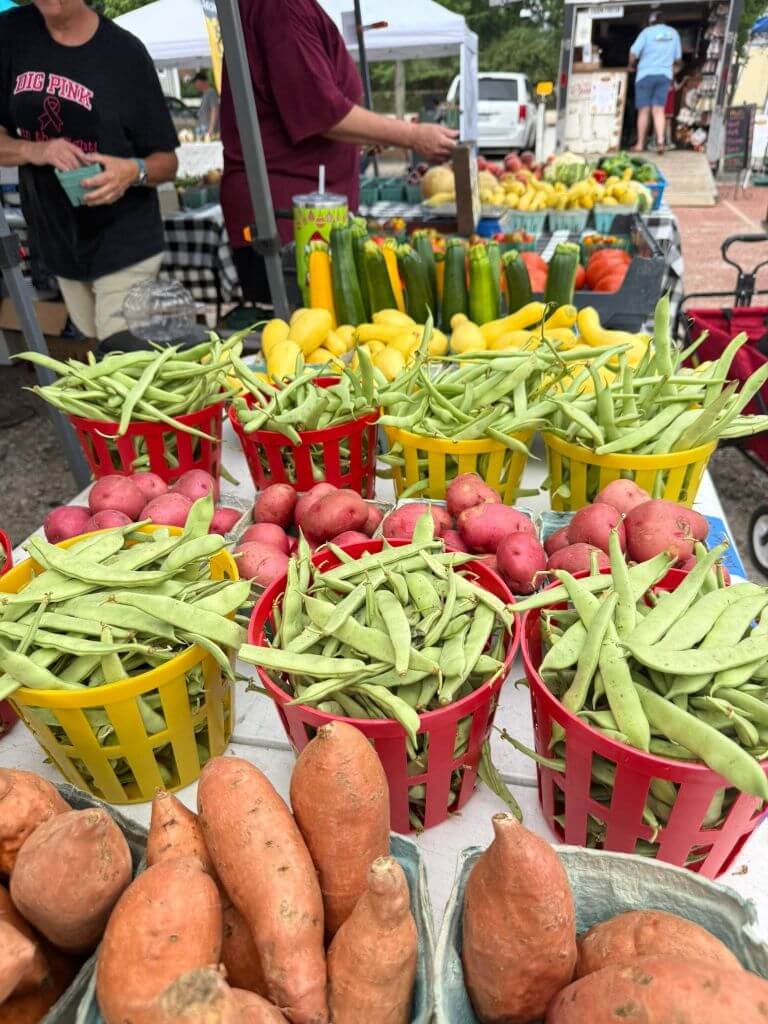 kcfmweb2 - Experience Camden, South Carolina Colorful baskets of green beans, red potatoes, and sweet potatoes are displayed on a table at an outdoor farmers market, with people and other fresh produce in the background.