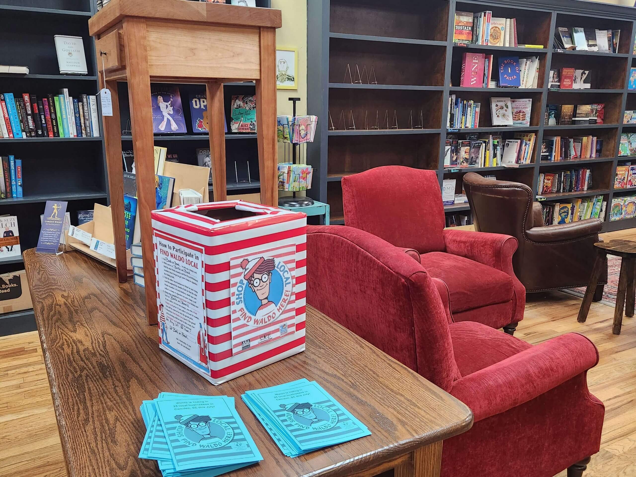 waldo1 A cozy bookstore features red chairs, wooden tables, and bookshelves. On a table in the foreground is a striped “Where’s Waldo?” box and blue flyers, inviting participation in a themed scavenger hunt.