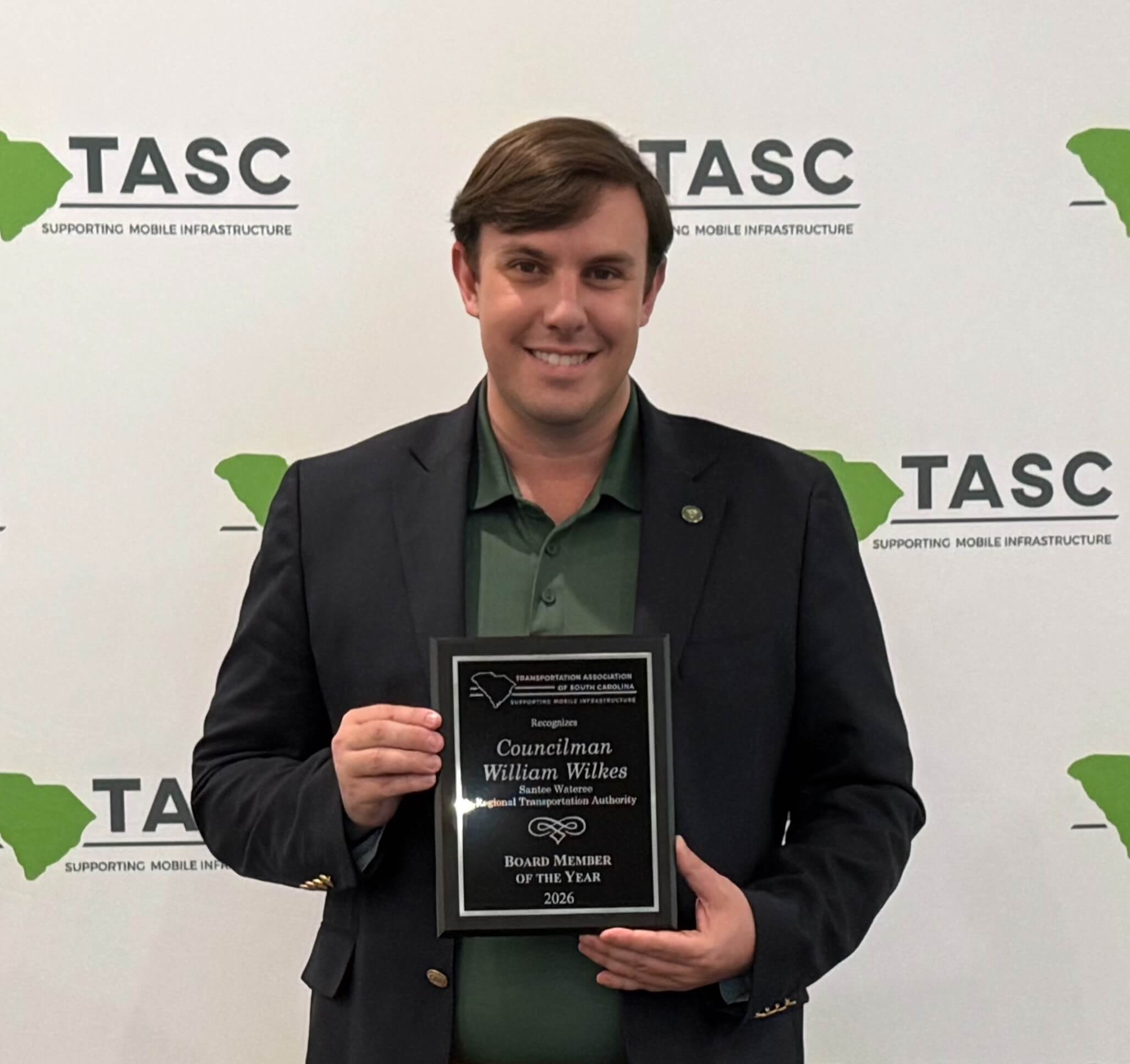 IMG_7786 A man in a green shirt and dark jacket holds a plaque award and smiles in front of a TASC-branded backdrop. The plaque recognizes him as Board Member of the Year for 2022.