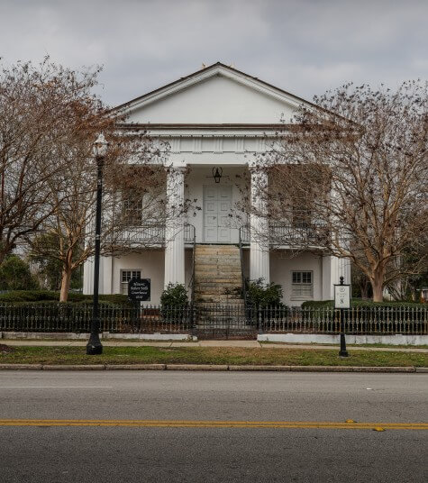 RMCH photo - Experience Camden, South Carolina A white, two-story historic building with columns and a staircase, surrounded by leafless trees and a black iron fence, sits across an empty street under a cloudy sky.