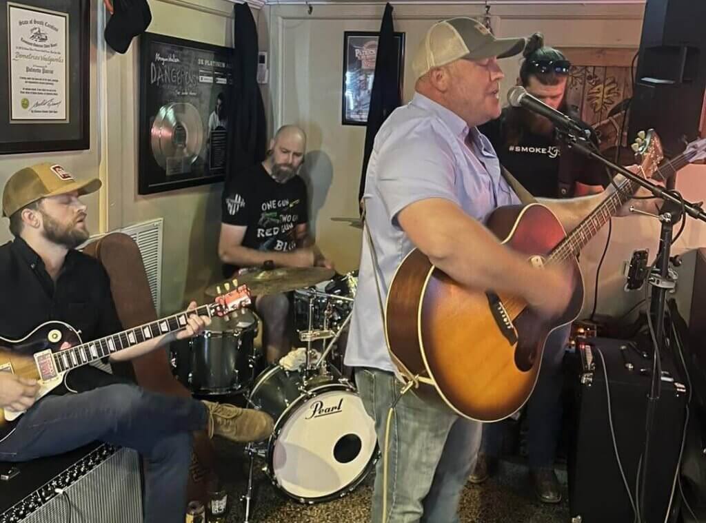 canebrake crop steeple - Experience Camden, South Carolina Four men play music in a small room: one sings with an acoustic guitar, one plays electric guitar, one is on drums, and one stands behind them with a bass guitar. Framed pictures and posters decorate the walls.
