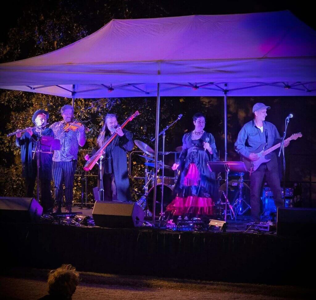 southerngothicband - Experience Camden, South Carolina A band of five musicians performs on an outdoor stage under a white canopy at night, lit by colorful lights. The group includes a violinist, guitarist, bassist, drummer, and a singer in a red and black dress.