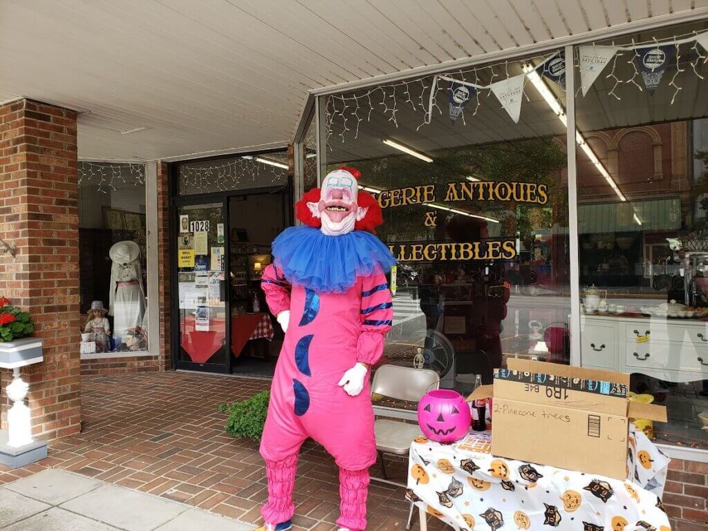 BooFest 2020 - Experience Camden, South Carolina A person in a bright pink clown costume with blue accents stands smiling outside an antique store, next to a table with a pink pumpkin bucket and various items on display.