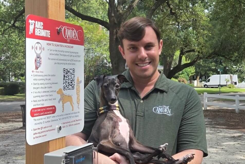 IMG_8017 A smiling man in a green shirt holds a small black dog next to a wooden post with pet safety and QR code signs, outdoors under trees.