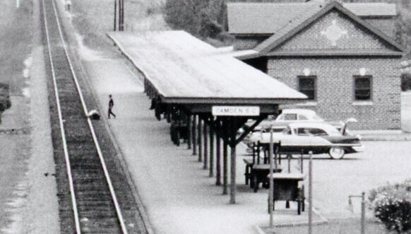 Seaboard Railroad Station.jpeg - Experience Camden, South Carolina Black and white photo of a small train station with one platform, a brick station building, vintage cars parked nearby, and railroad tracks stretching into the distance. A person walks along the platform.