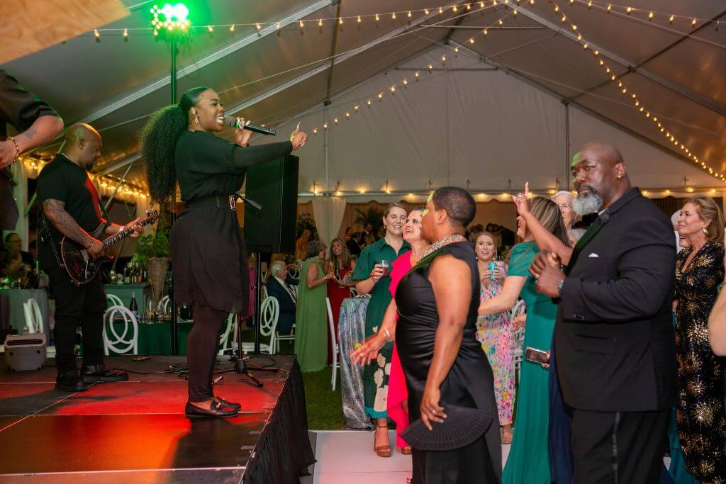 Starry Night Gala - Experience Camden, South Carolina A singer performs on stage at a lively indoor event as people dance and clap under a white tent decorated with string lights.