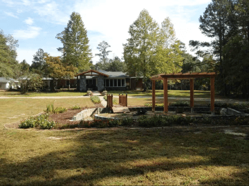 camden coach house - Experience Camden, South Carolina A large yard with a garden, pond, and wooden pergola in the foreground. A single-story house with large windows and surrounded by trees is visible in the background under a partly cloudy sky.