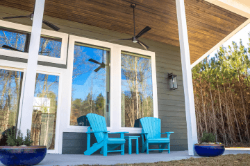 farmhouse at goat daddys - Experience Camden, South Carolina Two bright blue Adirondack chairs sit on a modern porch with large windows, ceiling fans, potted plants, and a partial view of trees reflecting in the glass.