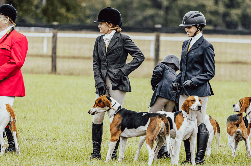 hounds17 - Experience Camden, South Carolina People in equestrian attire stand on grass with several hounds. Two women in black helmets and jackets are in focus, while another person in red stands to the left. A wooden fence runs along the background.
