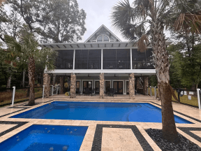 luxurylakehouse - Experience Camden, South Carolina A modern house with large windows and a screened porch overlooks a rectangular swimming pool and hot tub, surrounded by palm trees and stone tiles.