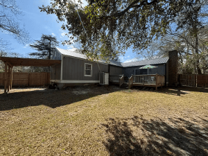 quiet cozy cottage - Experience Camden, South Carolina A single-story gray house with a fenced yard, small deck, and carport sits under a large tree on a sunny day.