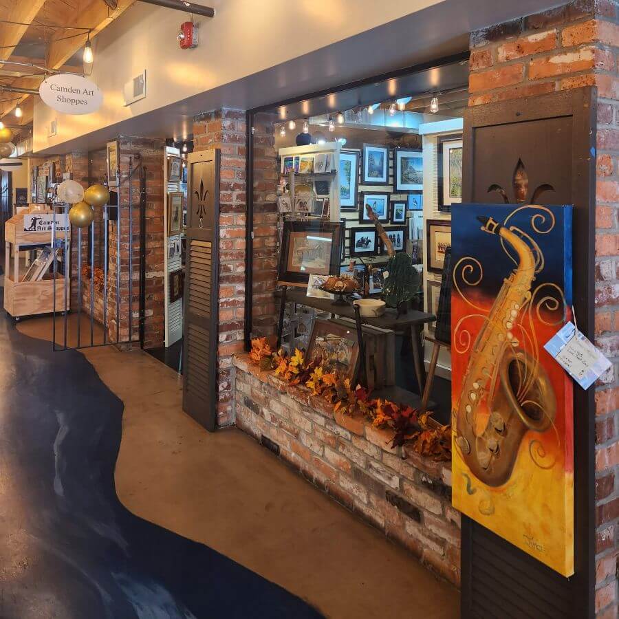 camdenartshoppes - Experience Camden, South Carolina A storefront for Camden Art Shoppes featuring a display window with framed artwork, autumn leaves, and a large painting of a saxophone on the brick exterior.