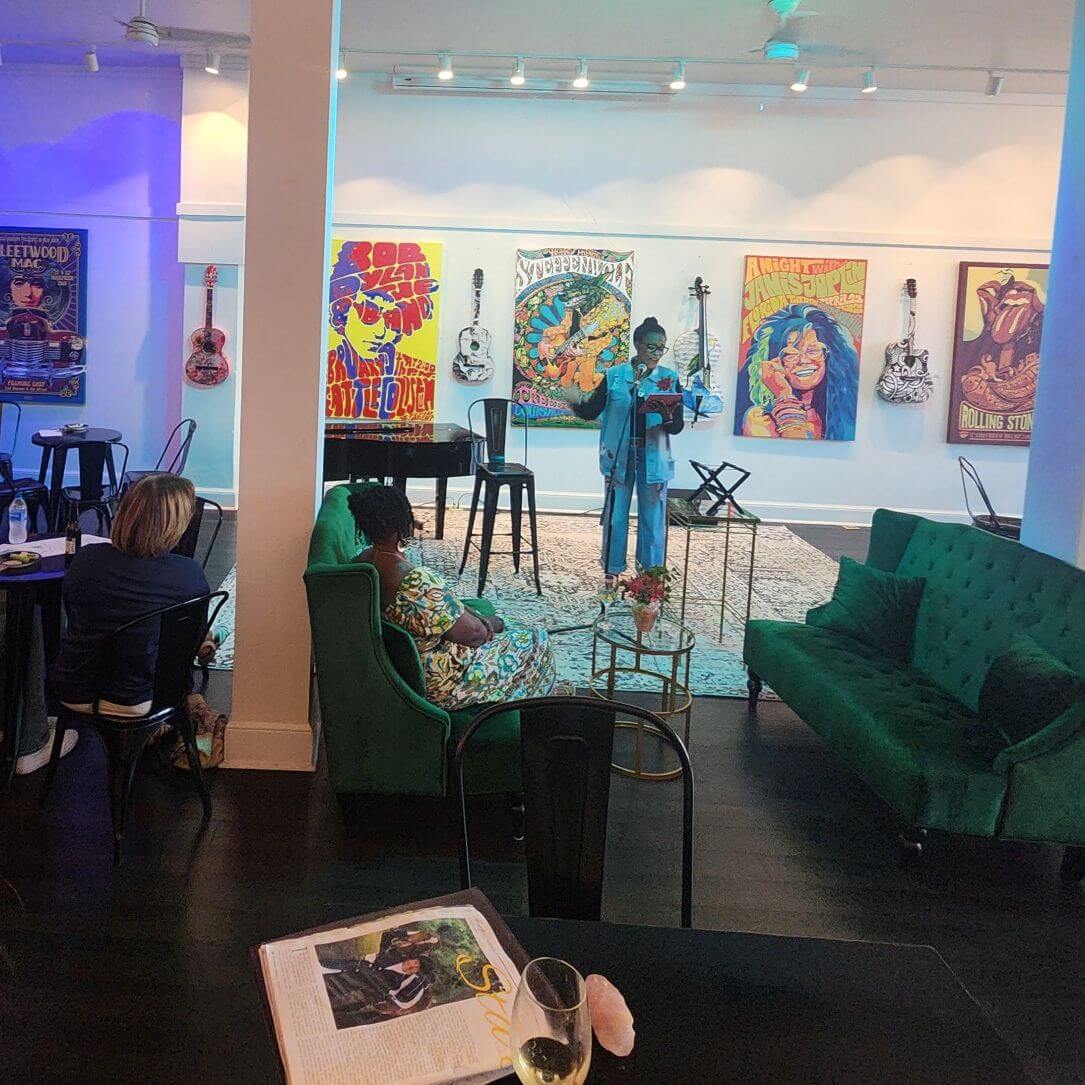 gallery537 - Experience Camden, South Carolina A person in a blue denim outfit stands in the Gallery 537 space reading from a book to a small audience, with colorful music posters and guitars displayed on the wall behind them.
